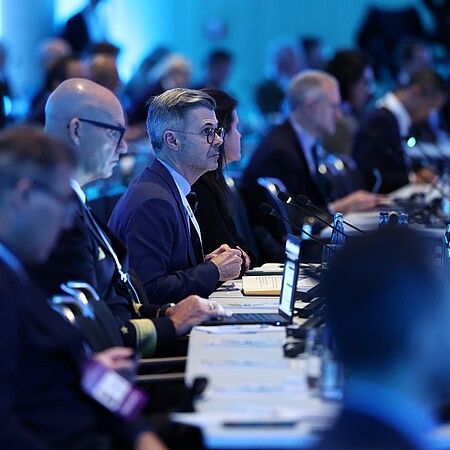 Reihe von Teilnehmenden bei einer Konferenz oder Sitzung, die an langen Tischen mit Mikrofonen und Laptops sitzen und aufmerksam nach vorne ausgerichtet sind; der Raum ist in kühles, blaues Licht getaucht, im Hintergrund sind weitere unscharfe Personen und Technik zu erkennen.