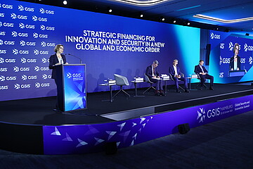 Conference stage at the GSIS (Global Security and Innovation Summit) in Hamburg. A speaker stands at a podium in front of a large blue LED screen displaying the title “Strategic Financing for Innovation and Security in a New Global and Economic Order.” Several participants are seated on stage as part of a panel discussion. A large screen on the right shows the speaker. The event takes place in a modern, professionally lit conference venue.