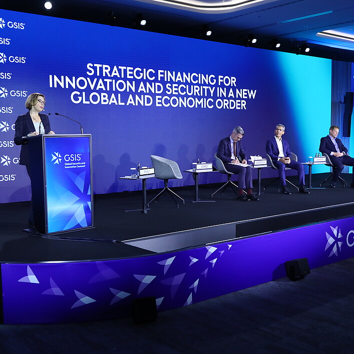 Conference stage at the GSIS (Global Security and Innovation Summit) in Hamburg. A speaker stands at a podium in front of a large blue LED screen displaying the title “Strategic Financing for Innovation and Security in a New Global and Economic Order.” Several participants are seated on stage as part of a panel discussion. A large screen on the right shows the speaker. The event takes place in a modern, professionally lit conference venue.