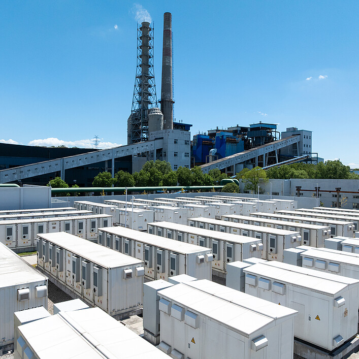 An areal view of rows of white battery energy storage systems under a clear blue sky with a large industrial building in the background.