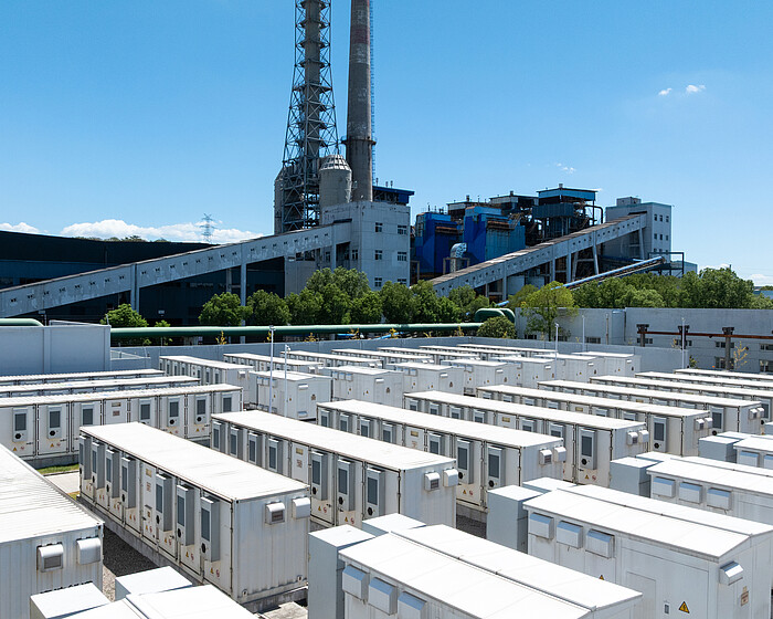 An aerial view of rows of white battery energy storage systems under a clear blue sky with a large industrial building in the background.