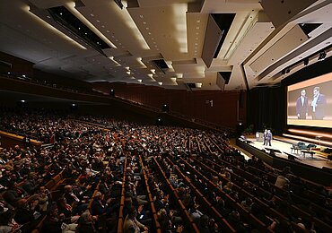 A fully packed auditorium with a modern ceiling. Two people are standing on a stage, and their images are projected onto a large screen in the background.