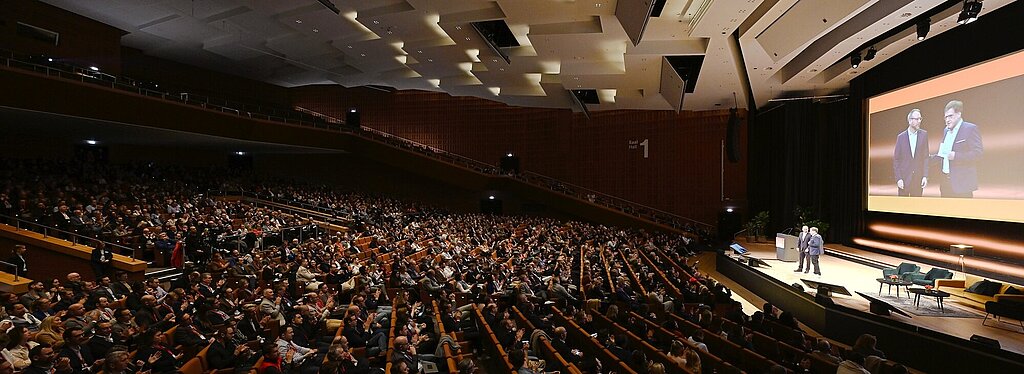 A fully packed auditorium with a modern ceiling. Two people are standing on a stage, and their images are projected onto a large screen in the background.