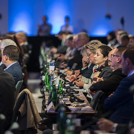 Teilnehmer einer Konferenz sitzen in Reihen an Tischen mit Mikrofonen und grünen Glasflaschen, während im Hintergrund Kameraleute das Geschehen dokumentieren. 