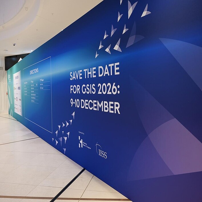 Große blaue Messewand in einem modernen Innenraum mit dem Schriftzug „Save the Date for GSIS 2026: 9–10 December“ sowie grafischen Dreiecksformen.